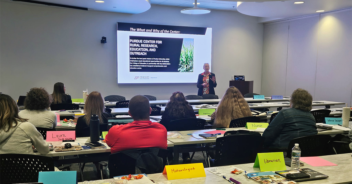 Jennifer Barce presenting in front of a classroom. Behind her is a presentation on the Purdue Center for Rural Research, Education, and Outreach.