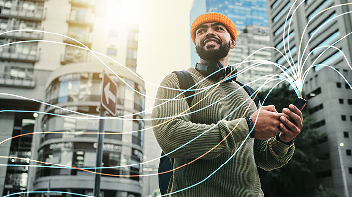 A black man with a beard stands on a city street holding a mobile device. There are digital lines superimposed on the image coming from the device.
