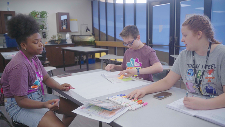 Three GEAR UP students seated at a table working on a poster. 