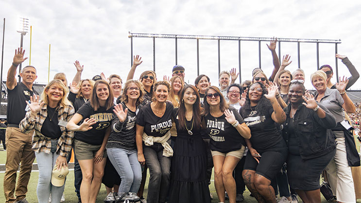 A group of Purdue community educators standing on a football field and wearing Purdue apparel. They are smiling and waving to the camera.