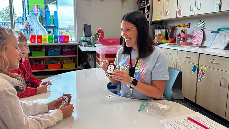 Kelsey (Sager) Rusk seated at a table in a classroom. She is smiling and teaching her students about numbers.