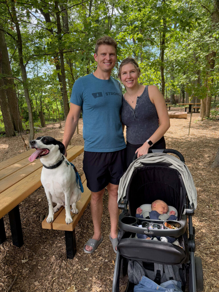 Isaiah Kent-Schneider standing with his wife, newborn baby, and dog in a nature trail. 