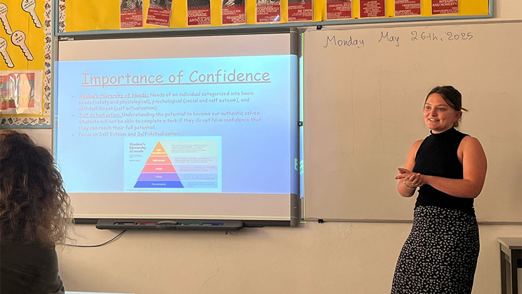 Sara Poulin standing at the front of a classroom. Projected next to her is a presentation with the currently slide titled, "Importance of Confidence".