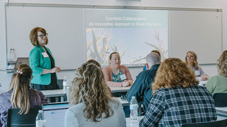 A presenter and group of panelists at the front of a classroom. Projected on a whiteboard is a presentation titled, "Cornfield Collaborations: An Innovative Approach to Rural Education".