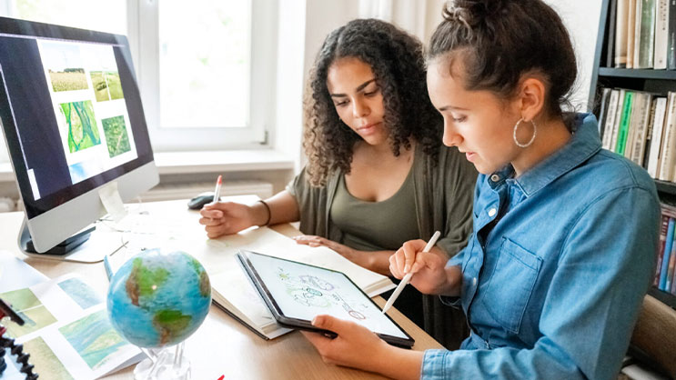 Two young women seated at a desk looking at a tablet. In front of them is a notebook, computer, and globe.