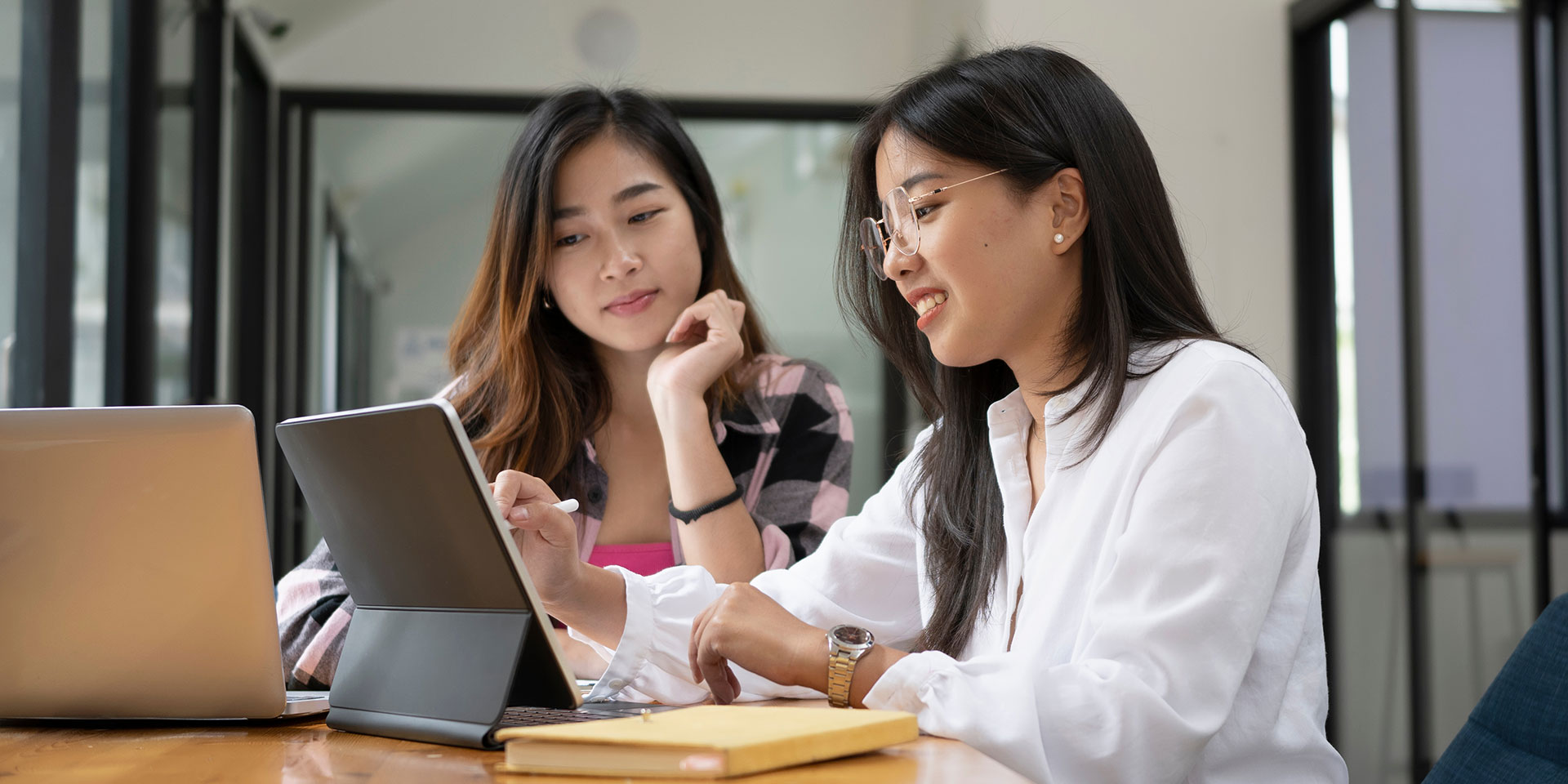 Two Asian women work on their laptops and tablets at a table in the library.