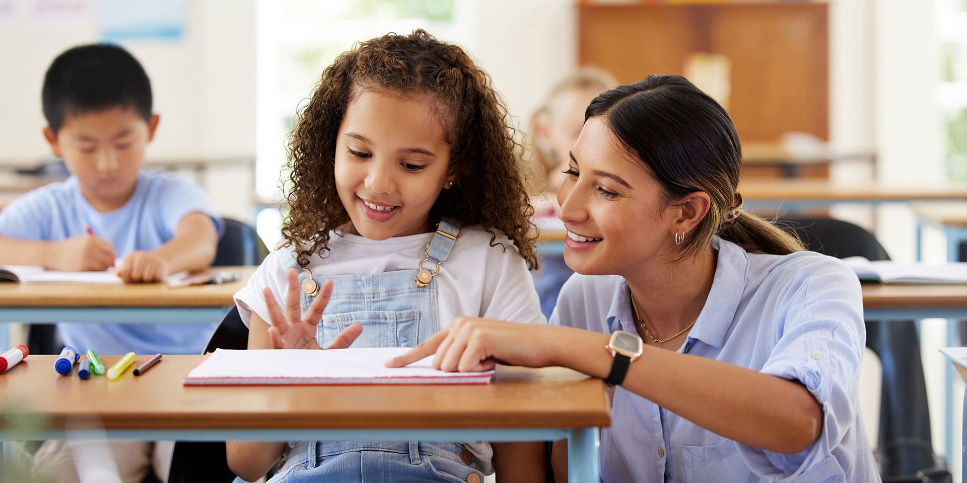 A teacher squats down and points at a notebook on a desk. Seated is an elementary school student.