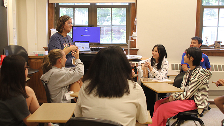 A teacher at the front of a classroom speaking to a small group of students.