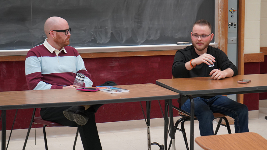 Ricky Price and Cooper Gretencord seated at a table at the front of a classroom. Ricky stares while Cooper speaks and gestures with his hands.