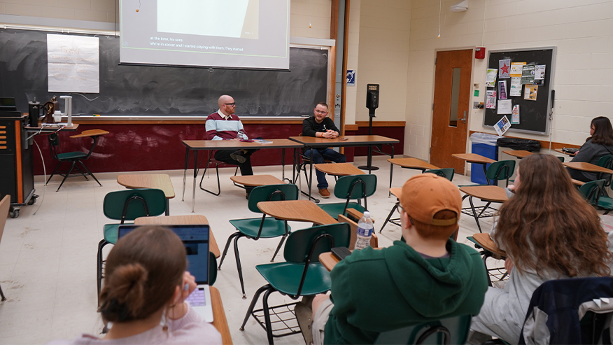 Ricky Price and Cooper Gretencord seated at the front of a classroom. A handful of students are seated in desks listening to the two men talk.
