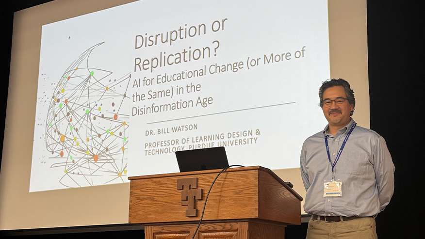 Bill Watson standing at a podium in front of a presentation titled, "Disruption or Replication? AI for Educational Change (or More of the Same) in the Disinformation Age".