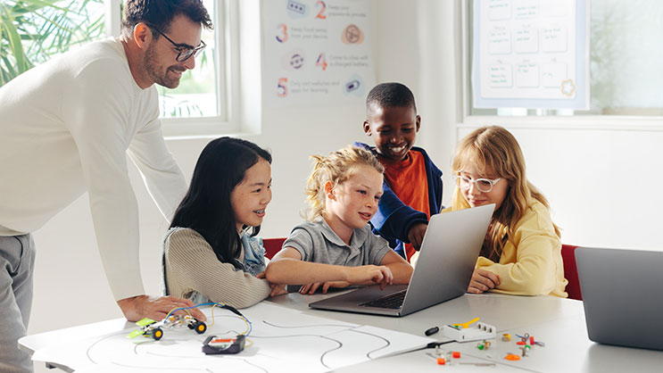 A group of students work on a robotics project using a laptop. A teacher watches them over their shoulders.