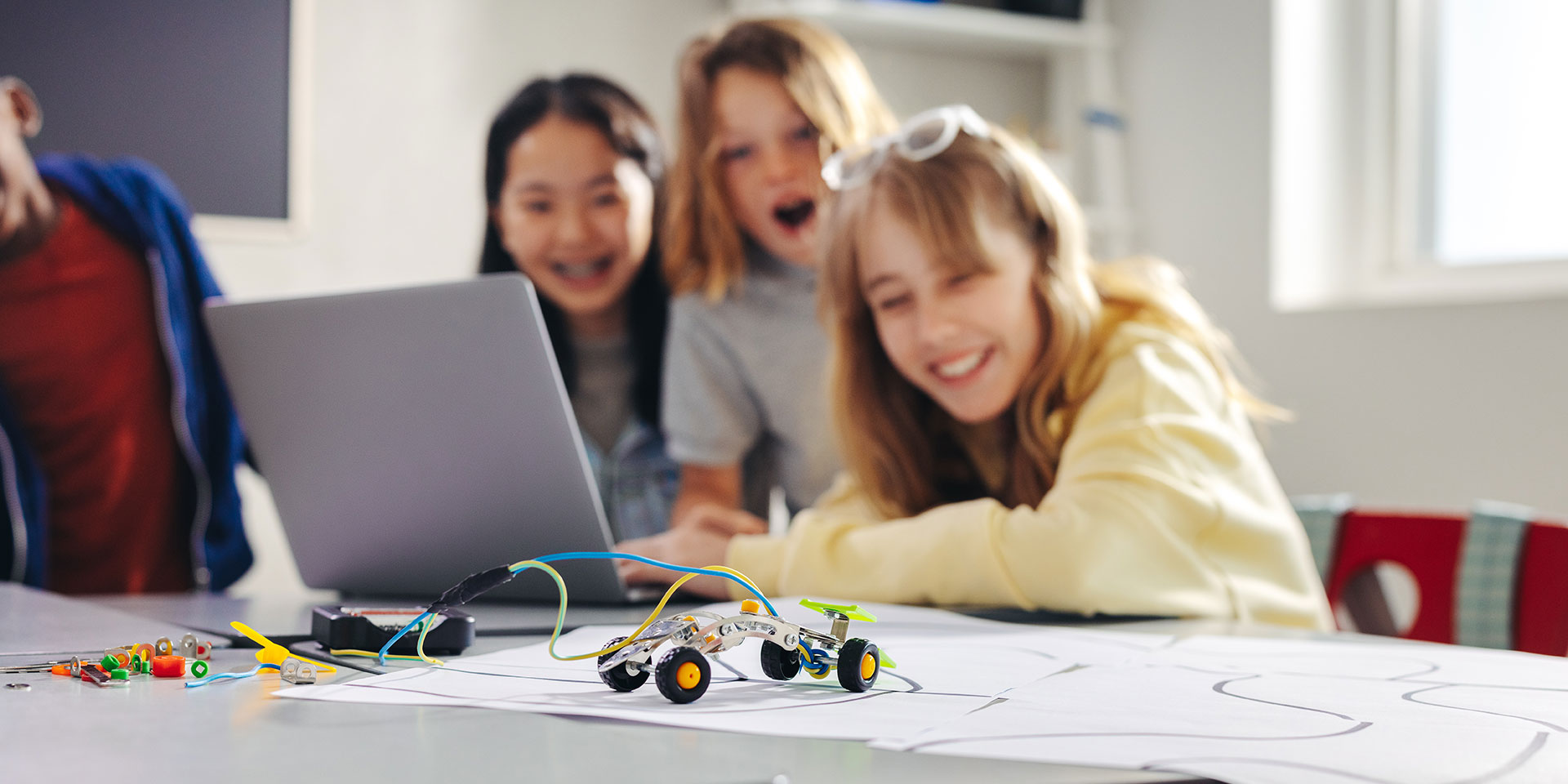 A robotic car is shown in the foreground while three students with delighted faces are blurred in the background
