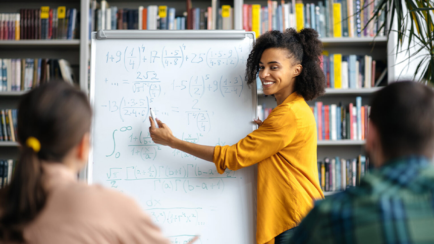 A teacher stands at a whiteboard pointing at math equations with a dry-erase marker.